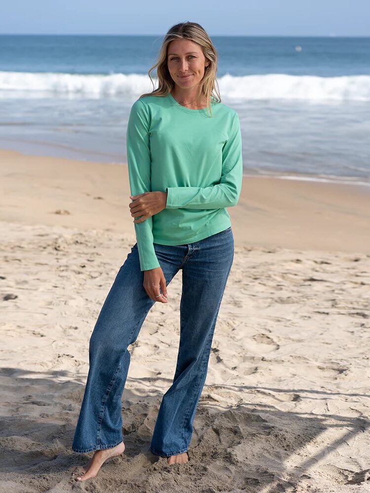 A woman in a green long-sleeve shirt and blue jeans stands barefoot on a sandy beach with waves in the background.