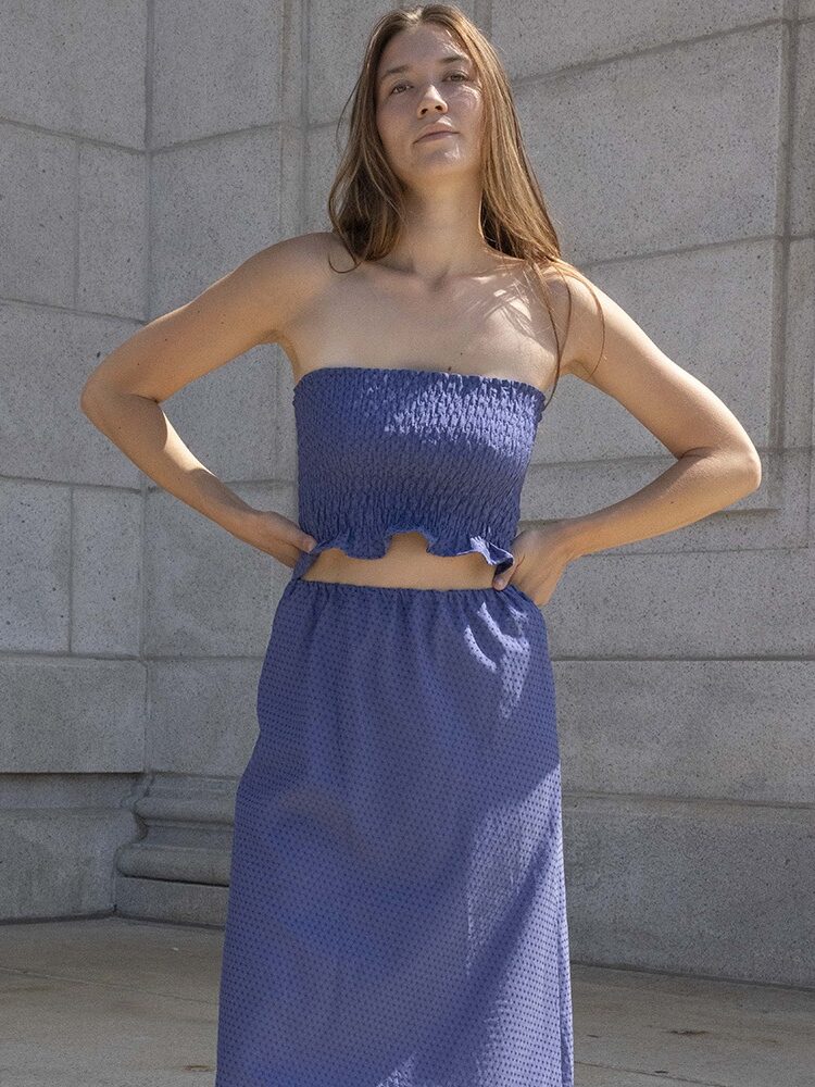 A woman stands against a stone wall, wearing a blue strapless top and matching skirt with a textured pattern.