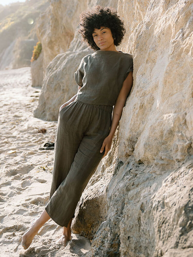 A person wearing a loose, olive-green jumpsuit leans barefoot against a rocky cliff on a sandy beach.