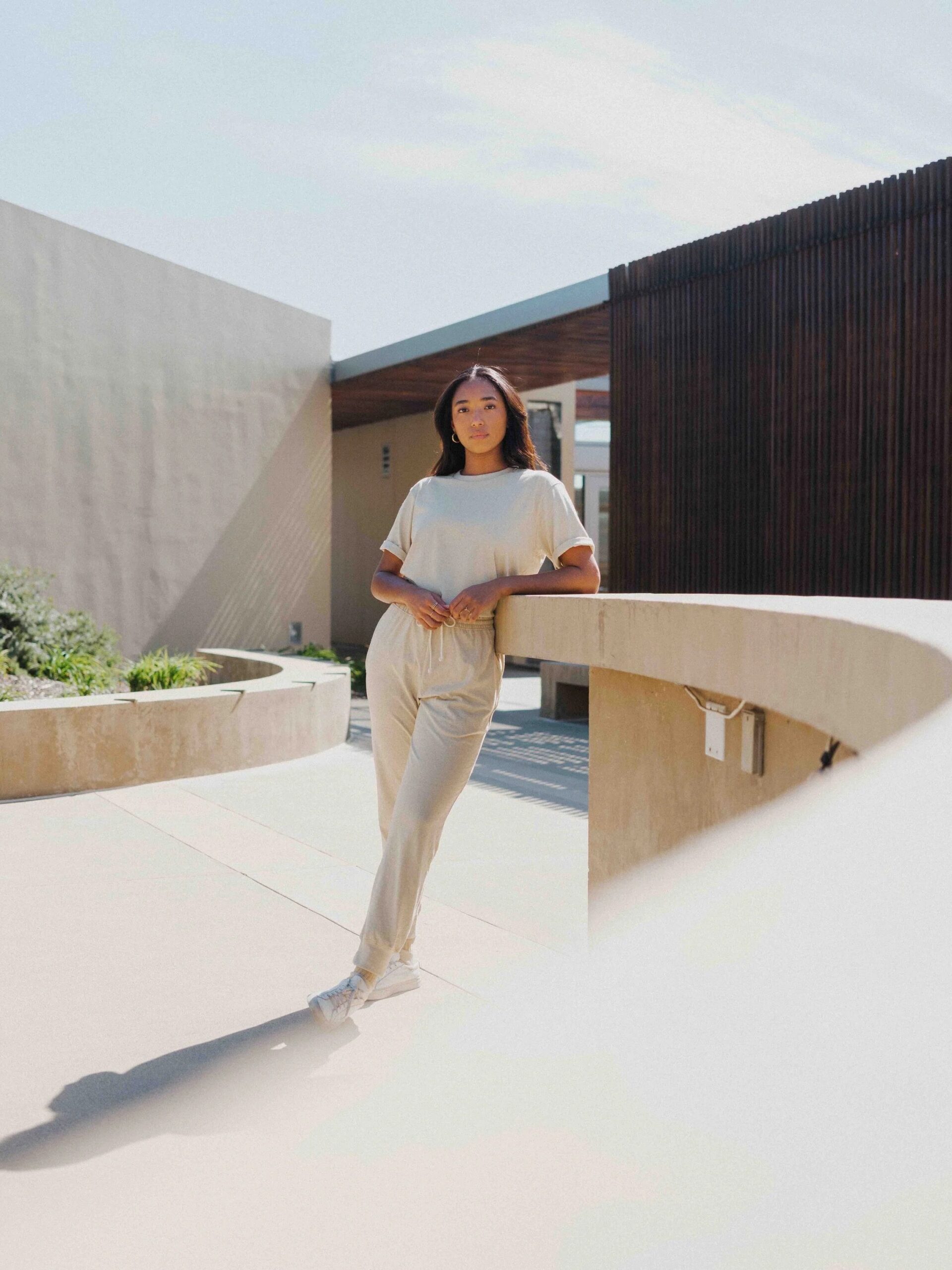 A woman in a light-colored outfit leans against a curved concrete railing in a modern outdoor setting with sunlight casting shadows.