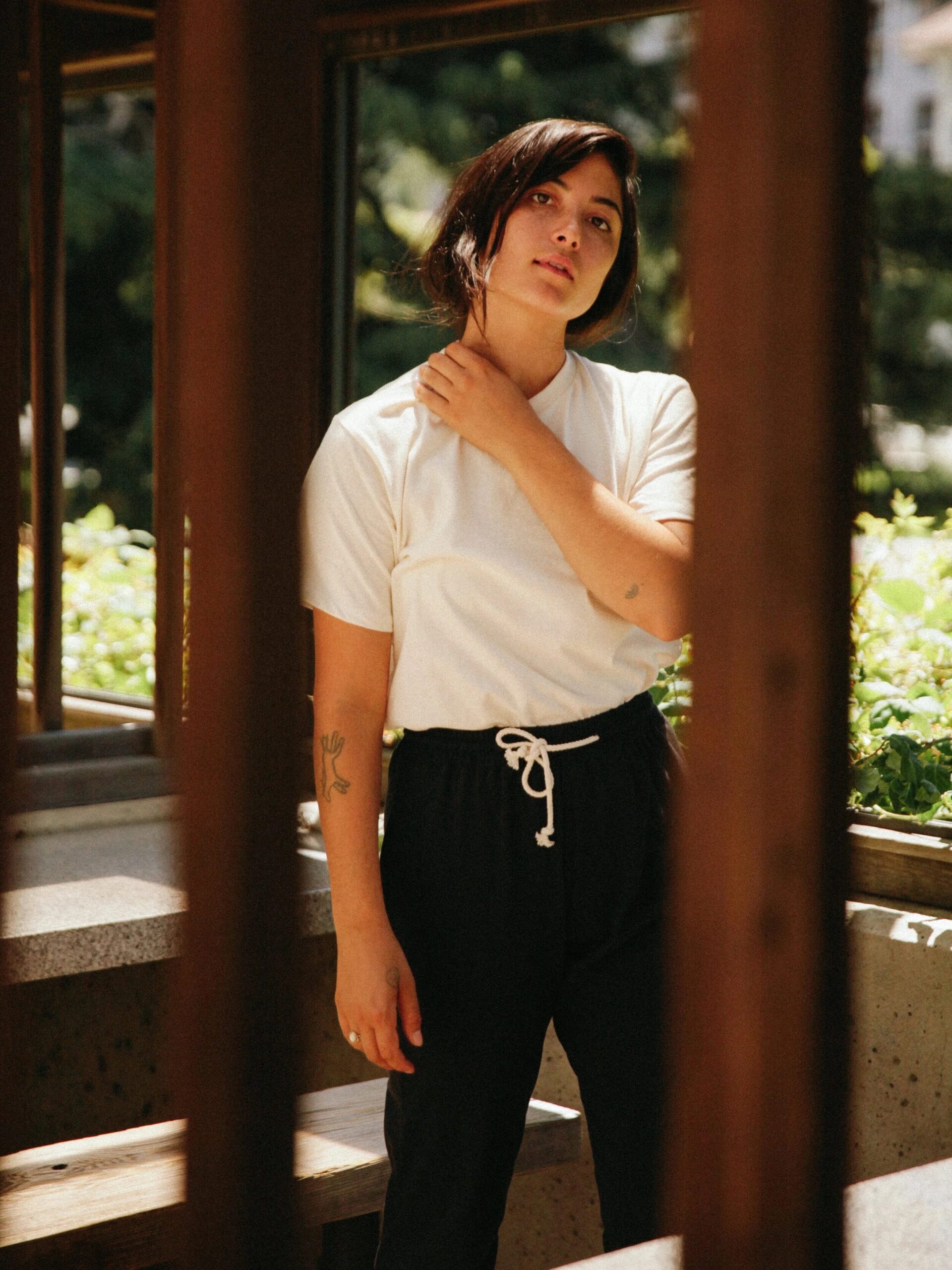 A person stands outdoors in a shaded area, wearing a white t-shirt and black pants, with one hand touching their neck and greenery visible in the background.