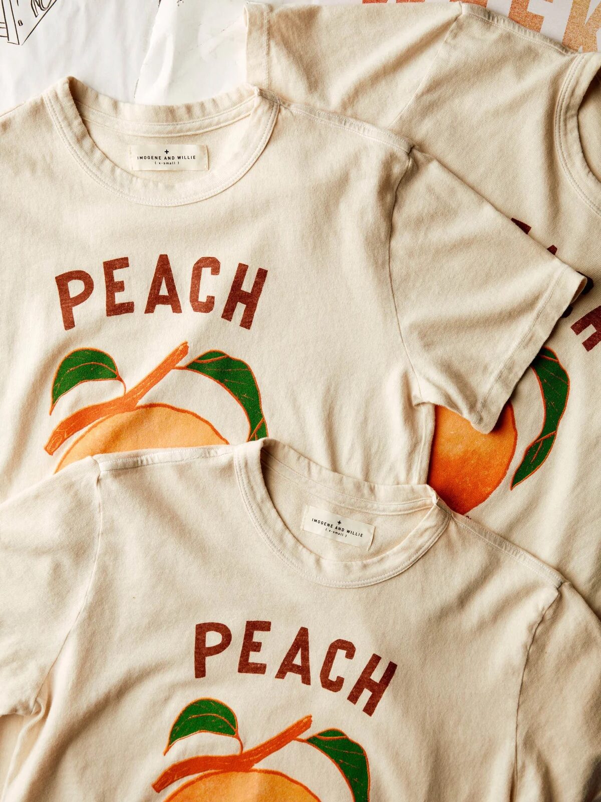 Three cream-colored t-shirts with "PEACH" text and a peach graphic are laid out overlapping each other.
