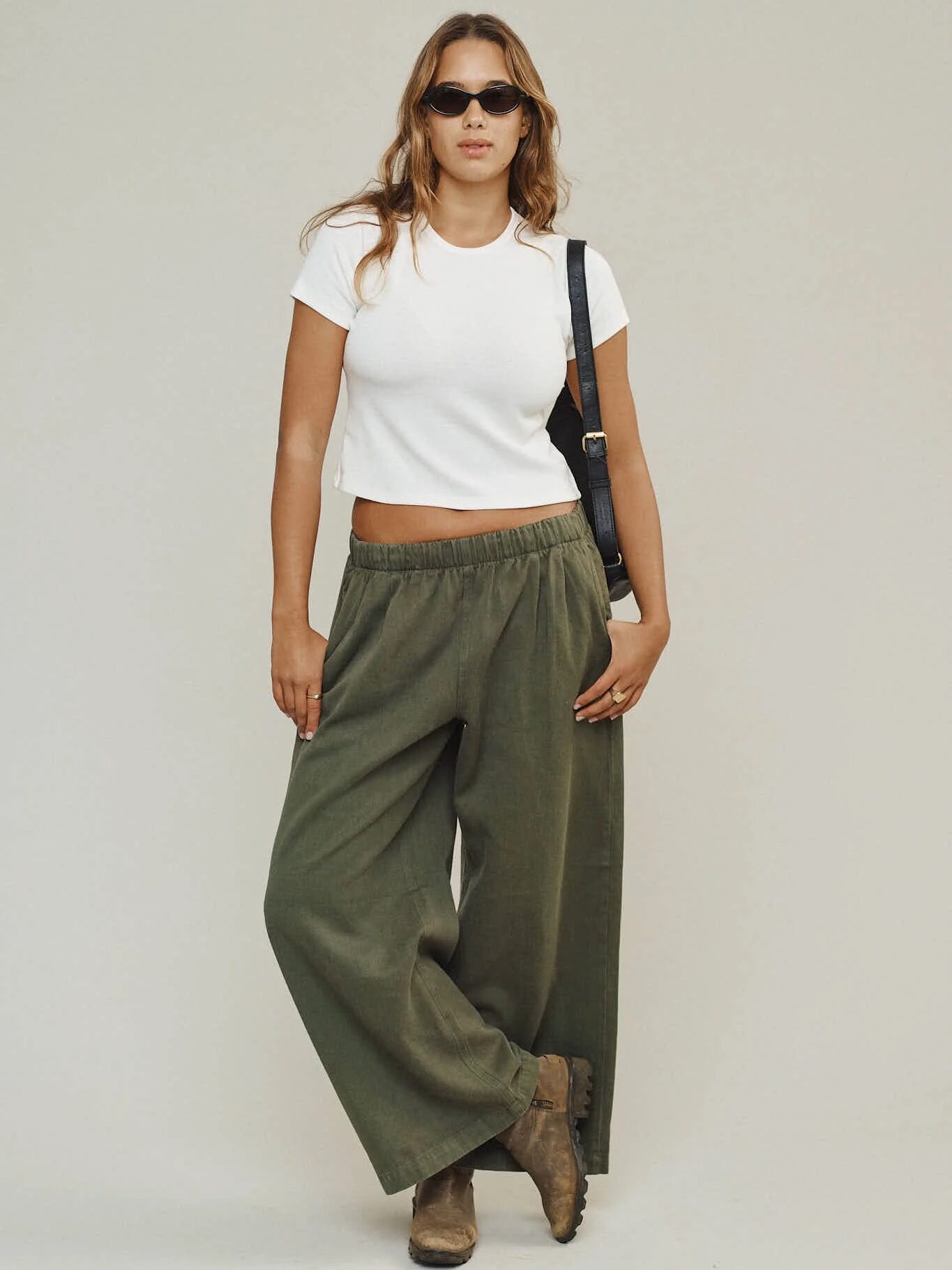 A person wearing a white cropped t-shirt, loose green pants, brown boots, sunglasses, and a black shoulder bag stands against a plain background.
