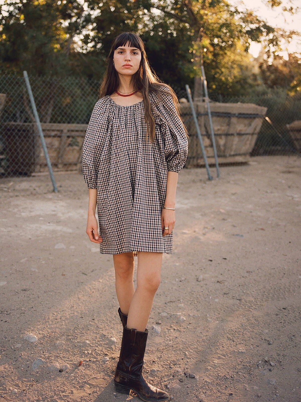 A woman in a checkered dress and black boots stands on a dirt ground outdoors, with a chain-link fence and trees in the background.