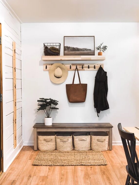 A tidy entryway with a black door, a bench with baskets, coat hooks holding hats and bags, a desk with a computer, a guitar on the wall, and decorative plants.