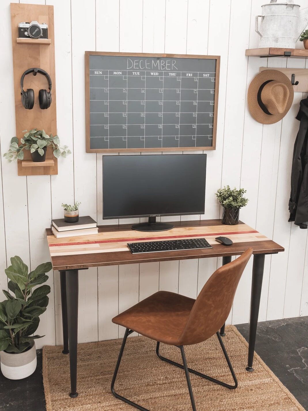 A minimalist workspace with a computer monitor, keyboard, and mouse on a desk, a brown chair, wall calendar, and various plants and decor against a white wall.