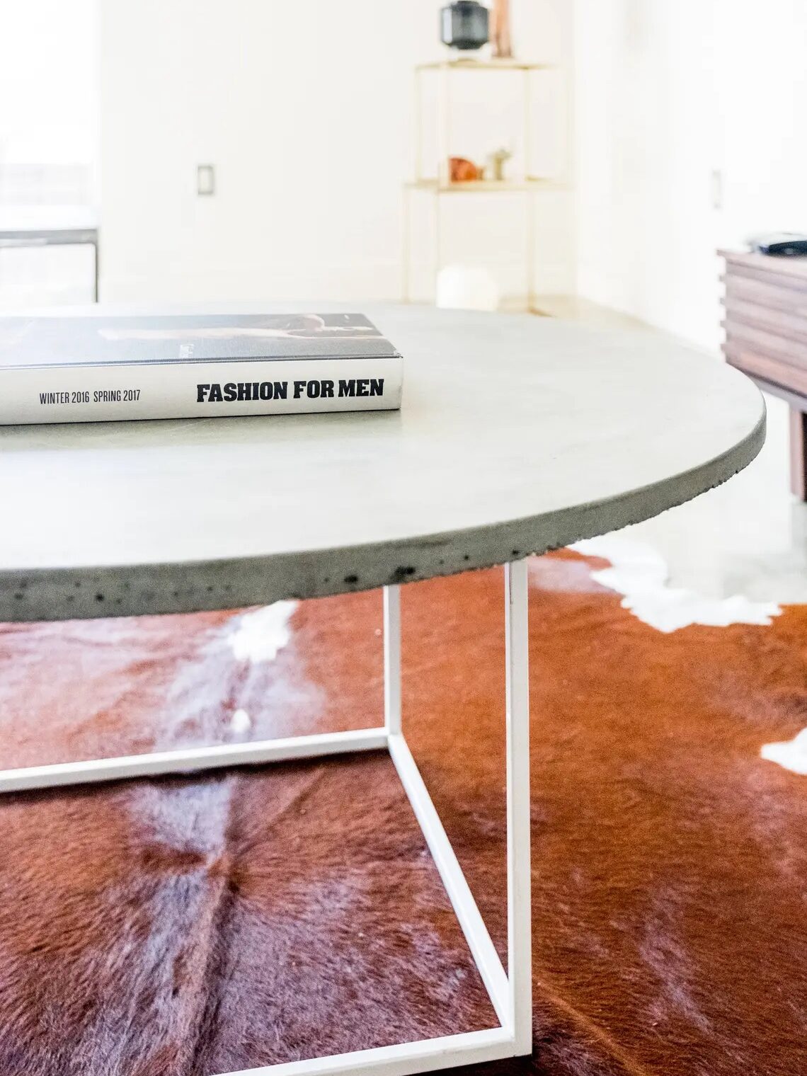A round concrete coffee table with a white metal frame holds a "Fashion for Men" book; a brown and white cowhide rug is beneath the table.