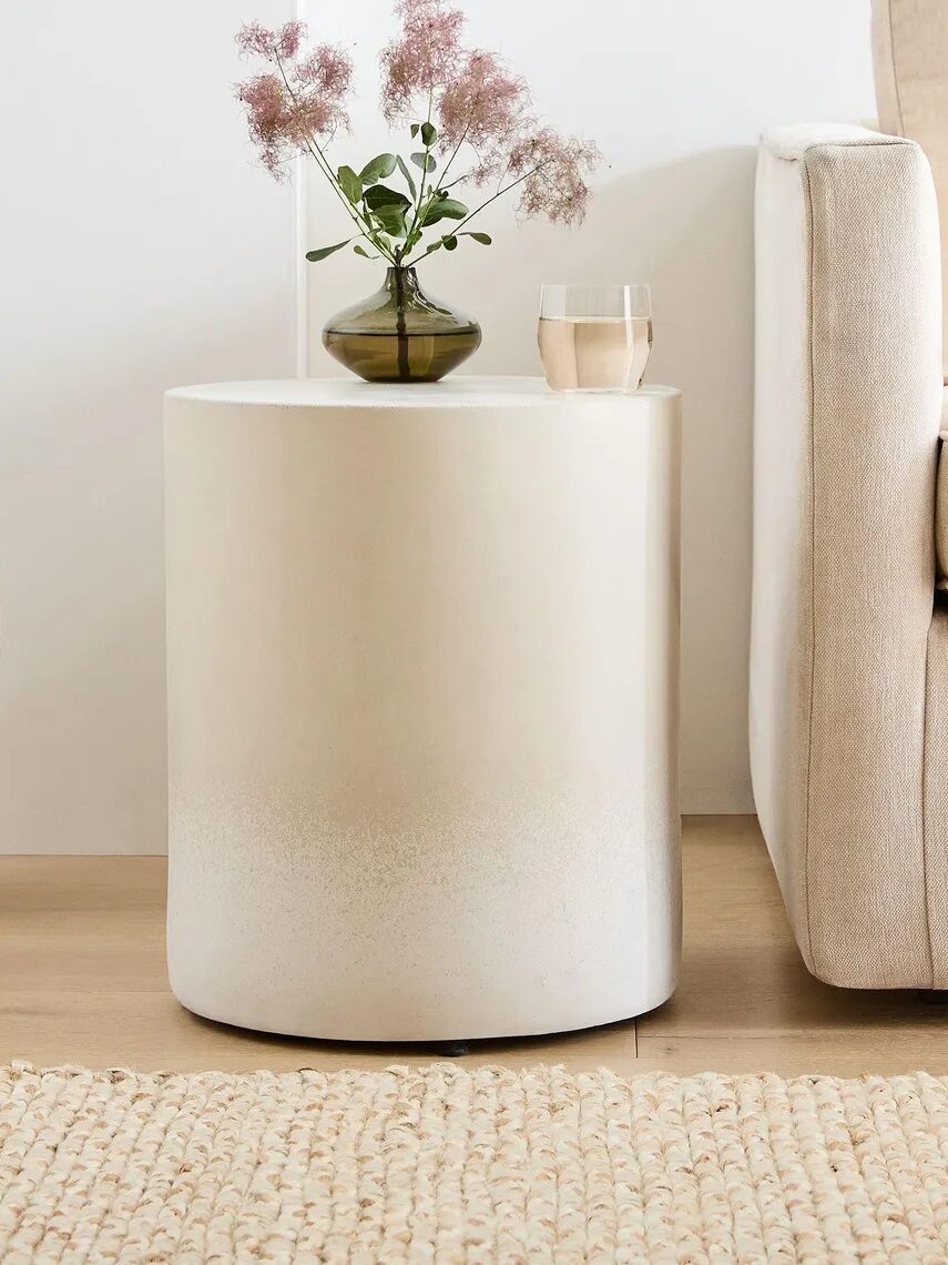 A round, white side table next to a beige sofa holds a small green vase with flowers and a glass of water. A textured rug and large vase are visible on the floor.