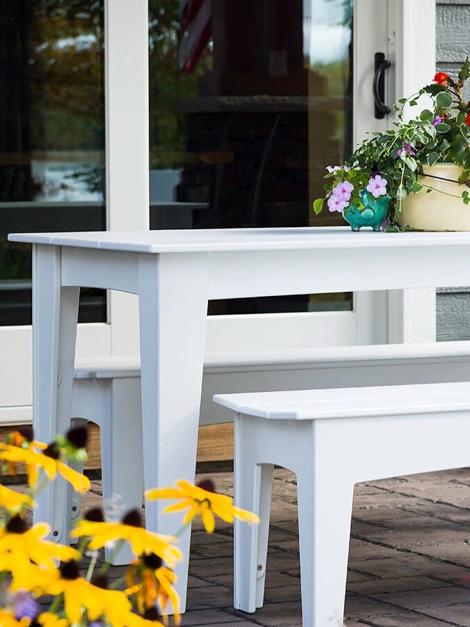 A white outdoor table with matching benches sits on a brick patio, decorated with a potted flower arrangement, surrounded by blooming yellow and purple flowers.