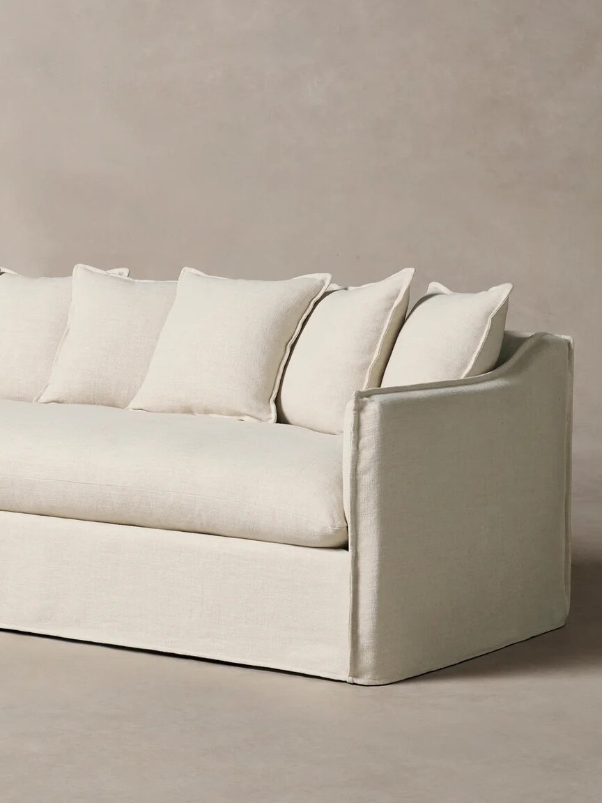 A modern, light beige sofa with a single seat cushion and seven matching back pillows, set against a neutral background.