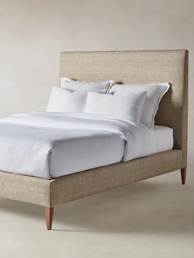 A beige upholstered bed with a tall headboard, white bedding, and wooden legs set against a plain neutral background.