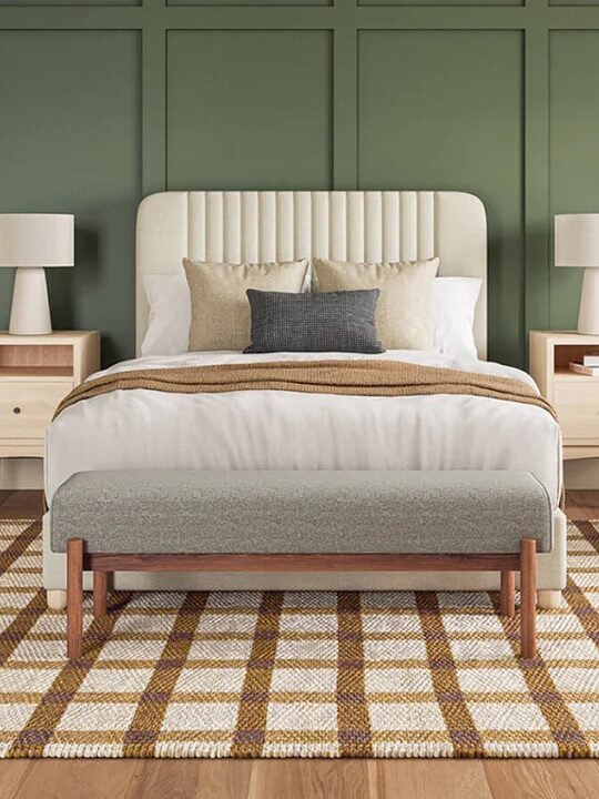 A modern bedroom with a green paneled accent wall, upholstered bed, beige armchair, wooden furniture, and a plaid area rug.