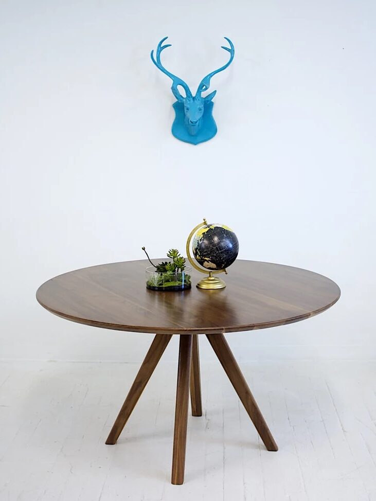 A round wooden table with a small globe, a potted succulent, and a pen rests against a white wall with a blue deer head sculpture mounted above.