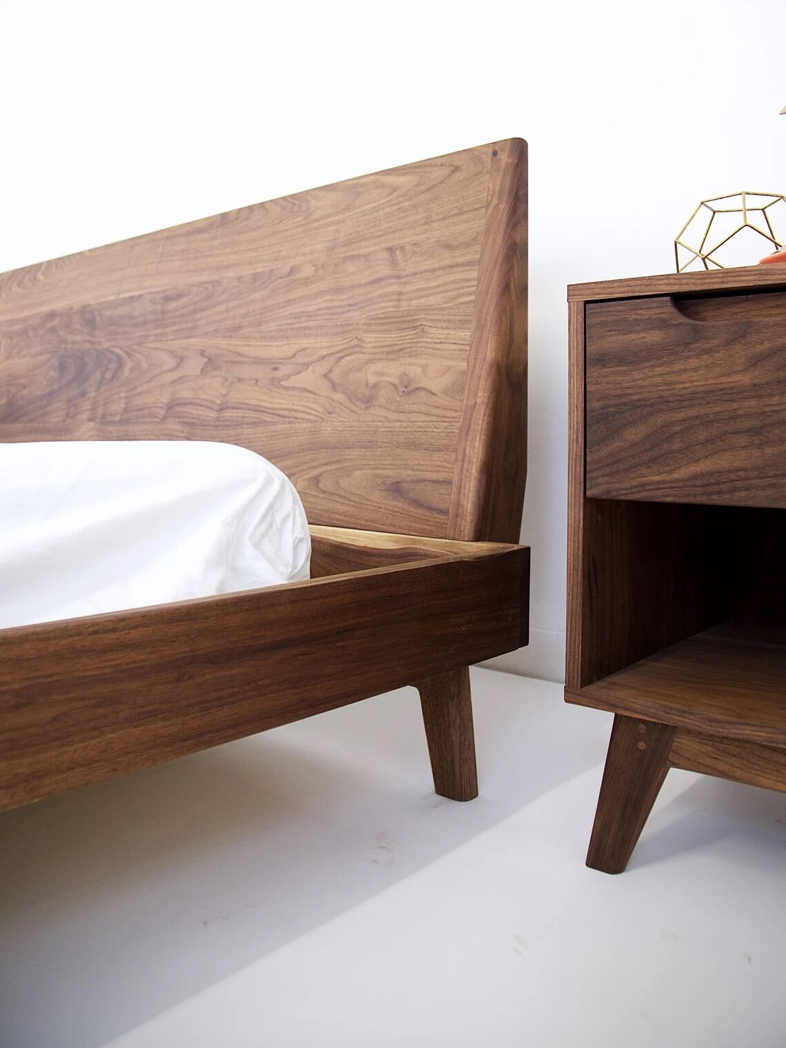 A wooden bed frame with a slanted headboard next to a matching nightstand, both with a mid-century modern design, set against a white background.