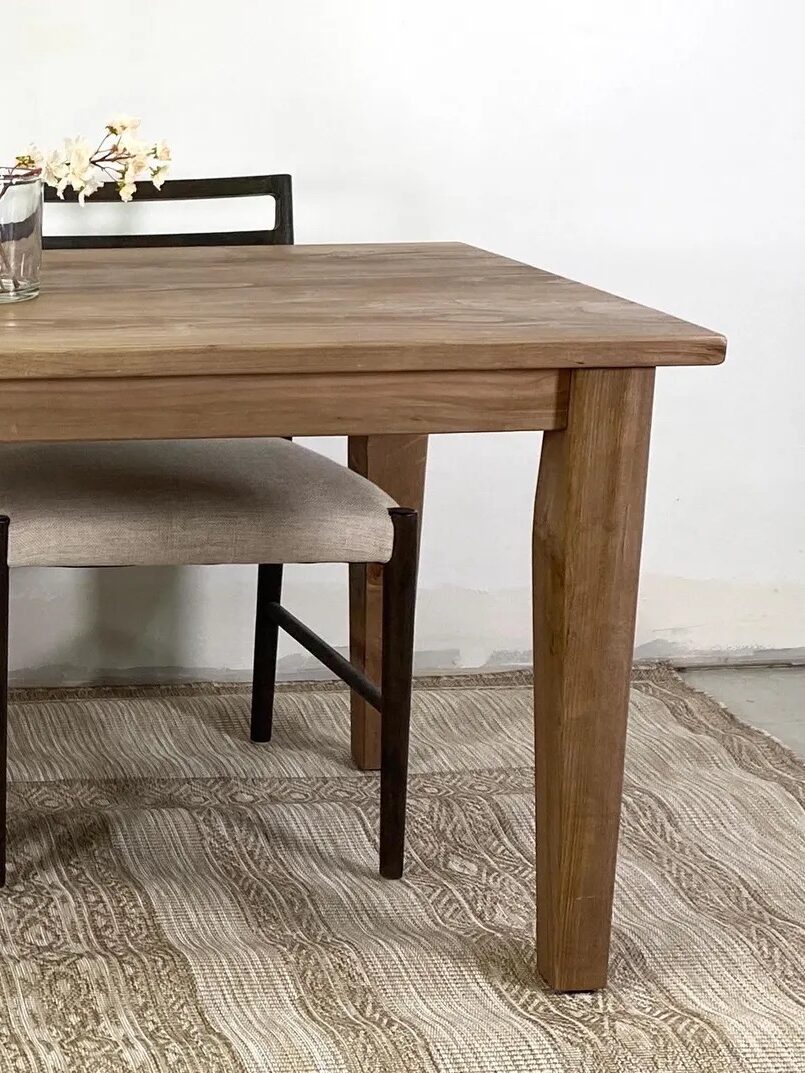 A wooden dining table with two cushioned chairs, a glass vase with flowers on top, set on a patterned rug against a plain wall.