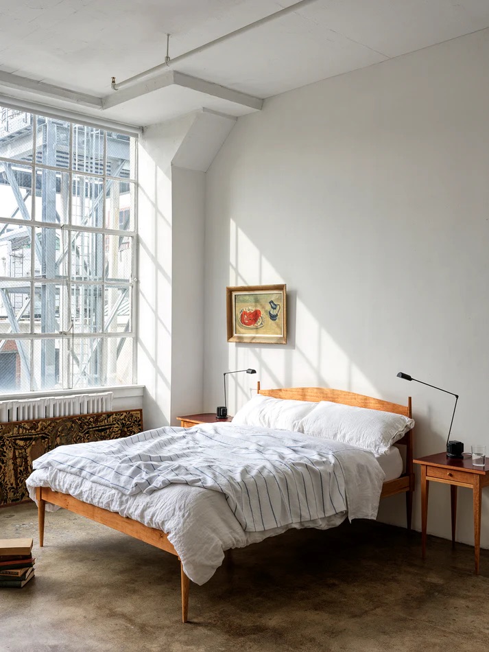 A minimalist bedroom with a wooden bed, white bedding, two small side tables, reading lamps, a framed painting, and large windows letting in natural light.
