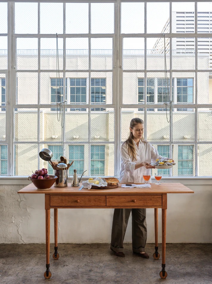 A woman stands behind a wooden table set with food and drinks in front of large industrial windows.
