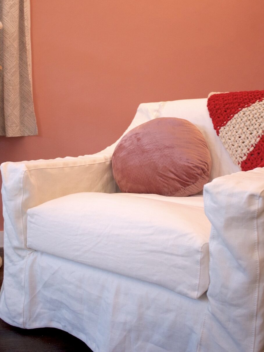 A white armchair with a pink round pillow and a red-and-white crocheted blanket sits against a pink wall.