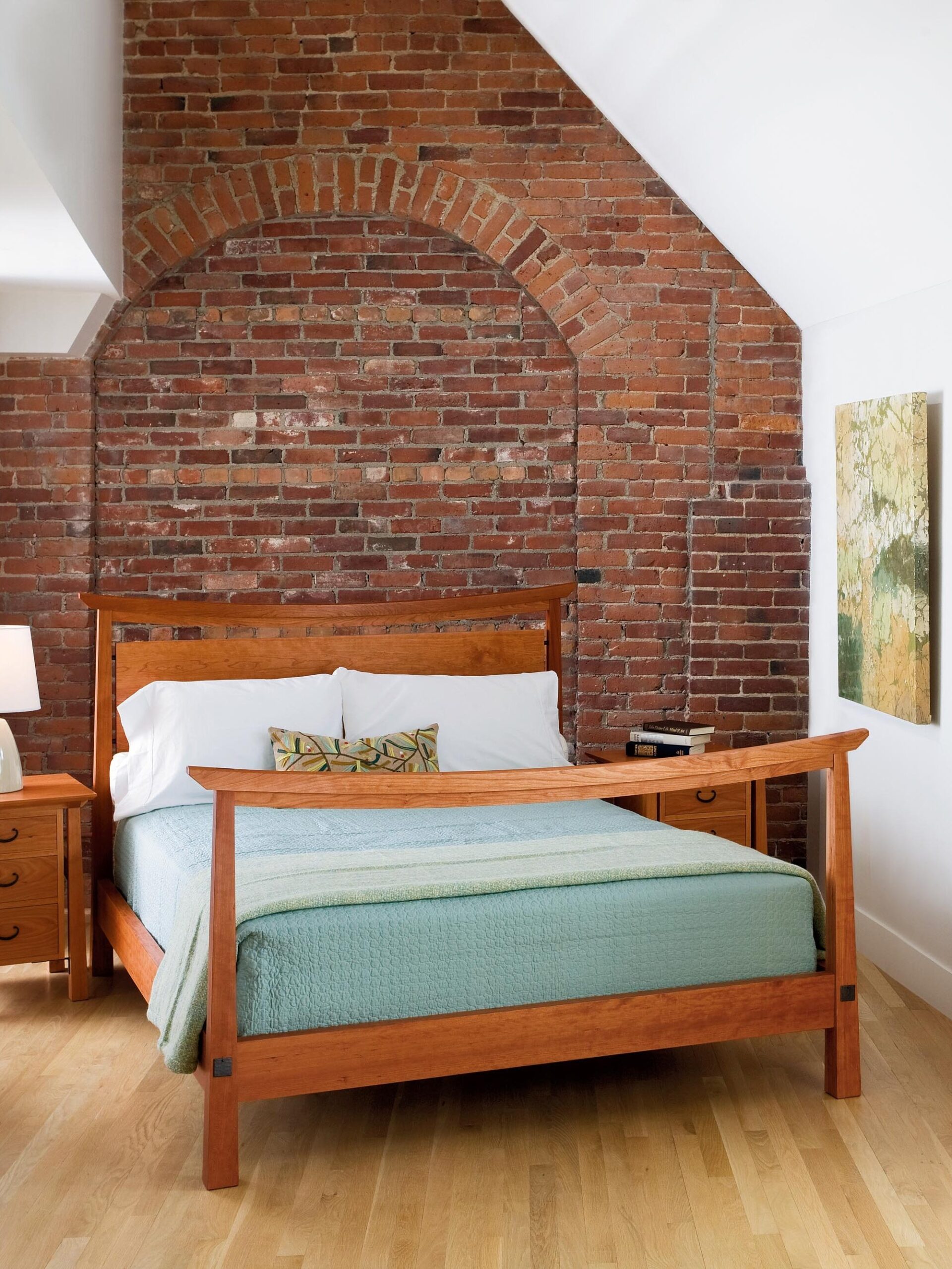 A bedroom with exposed brick walls, a wooden bed with a green quilt, a matching nightstand, a dresser, a table lamp, and a framed artwork on the wall.