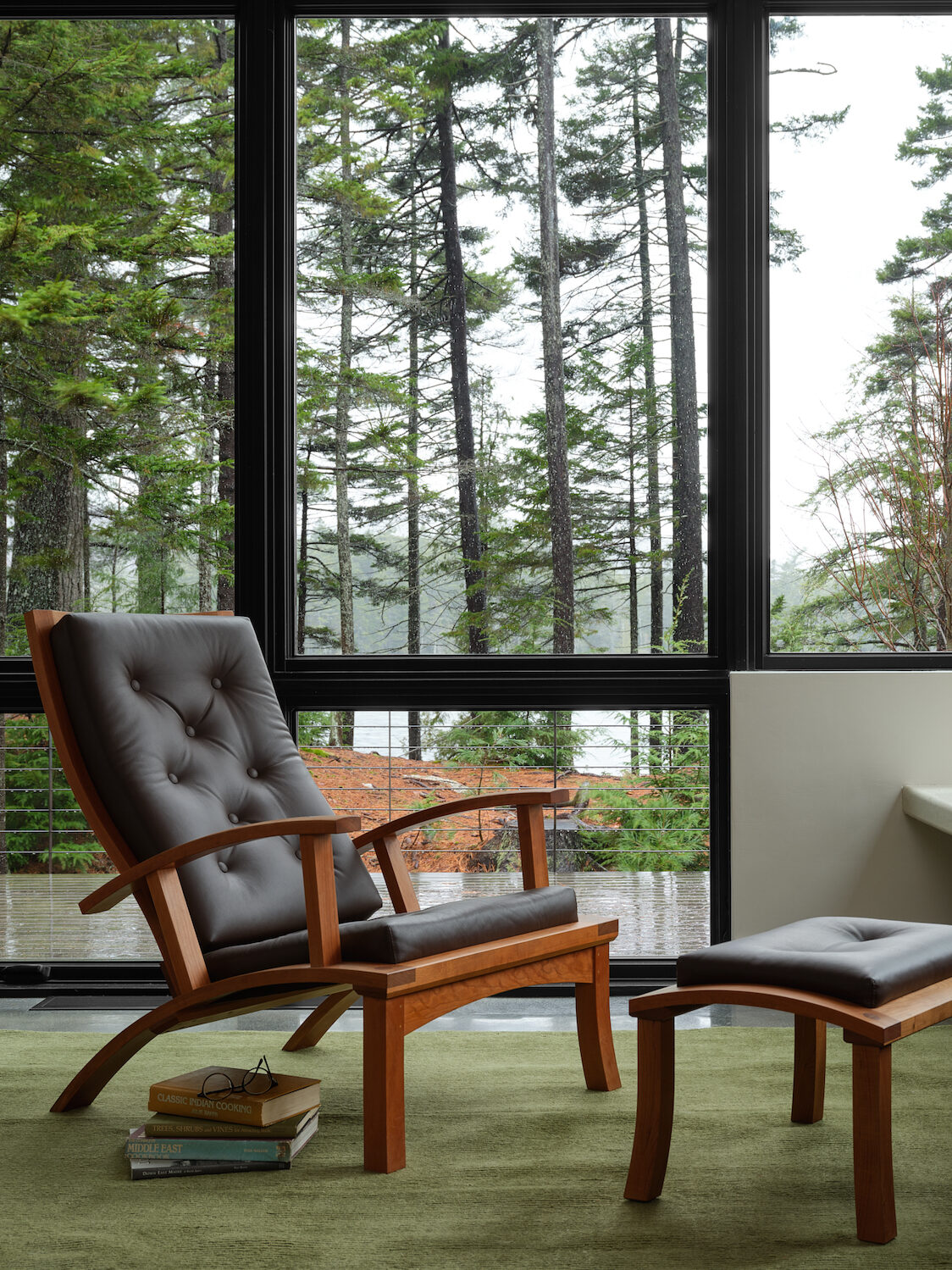A wooden armchair with a matching ottoman sits on a green rug by large windows overlooking a forest. A built-in bench with two woven baskets is against a tiled wall.