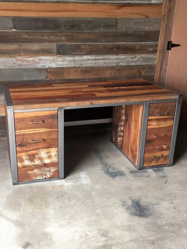 A wooden desk with metal frame and six drawers stands on a concrete floor against a wall made of horizontal wooden planks.