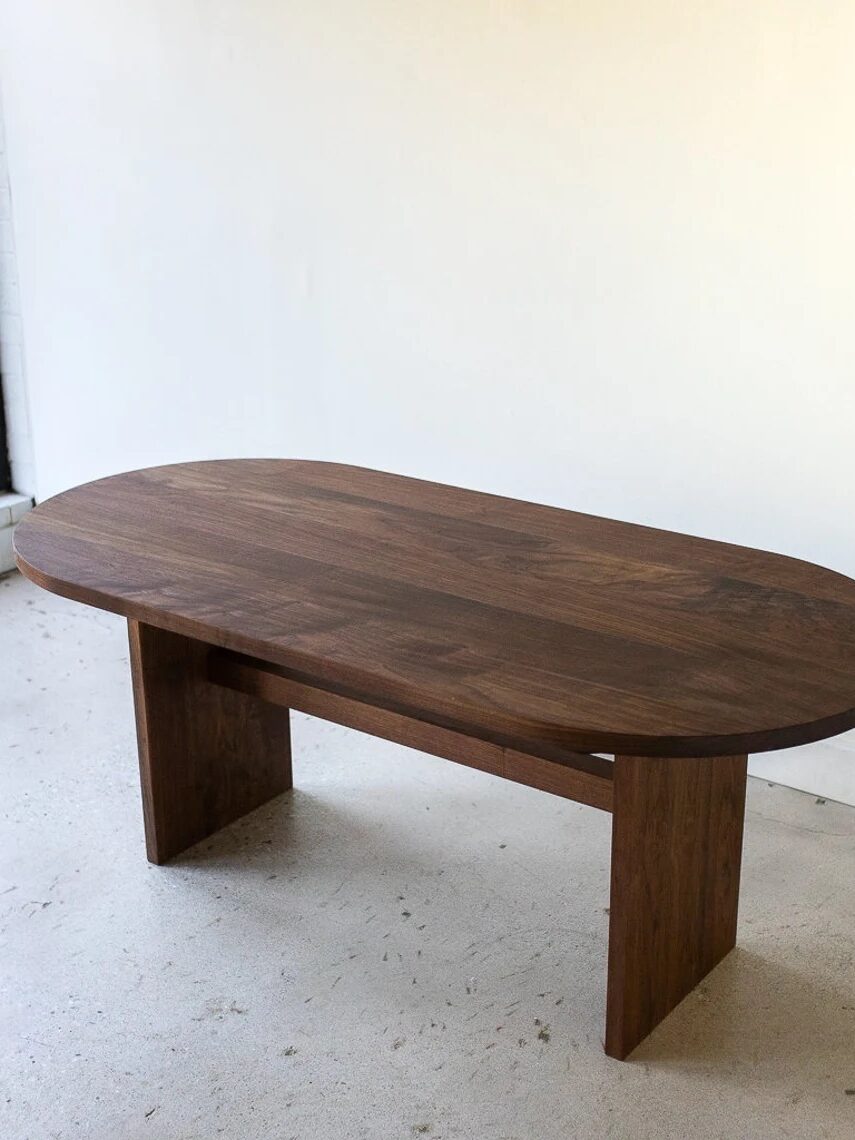 A dark wooden oval table with two wide legs stands in a minimally furnished room with white walls and floor near a window.