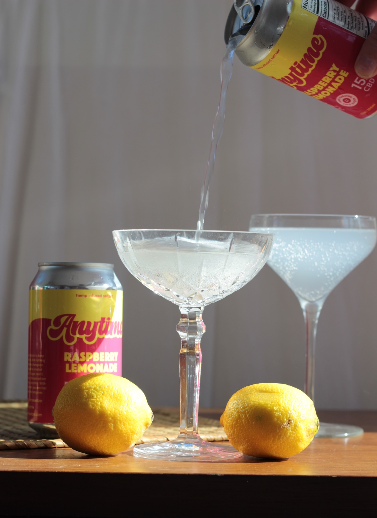 A can of Anytime Raspberry Lemonade is being poured into a coupe glass, with another can, two lemons, and two filled glasses on a wooden table.