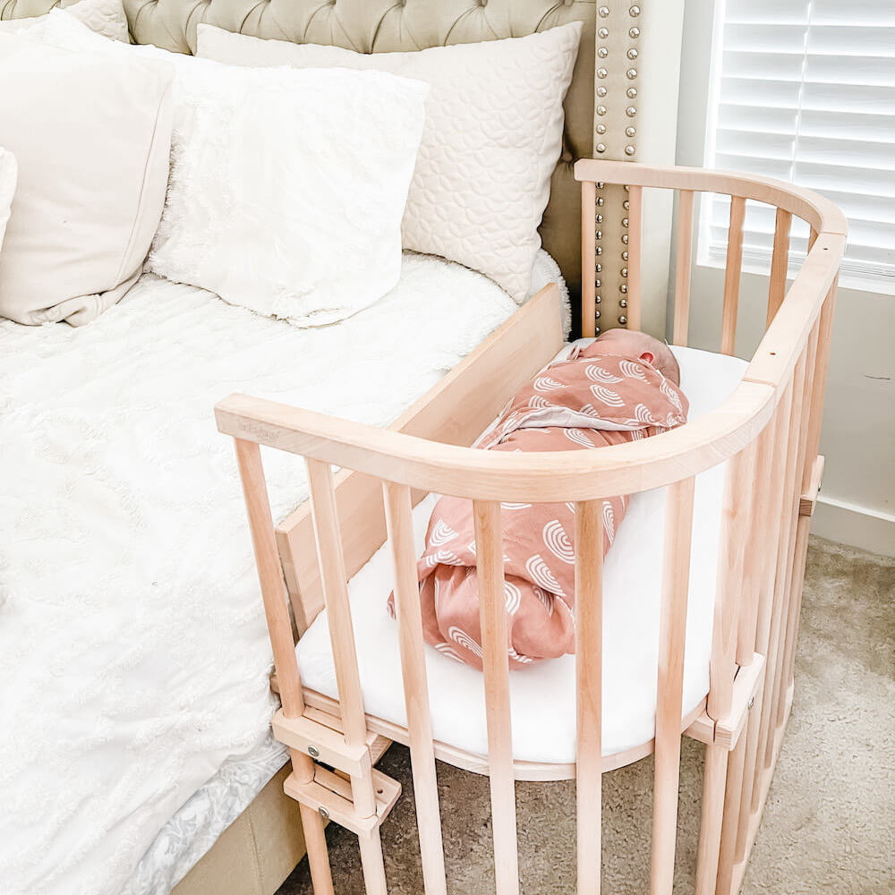 A wooden bedside bassinet attached to a bed contains a baby swaddled in a pink blanket with white patterns, next to a window with blinds.