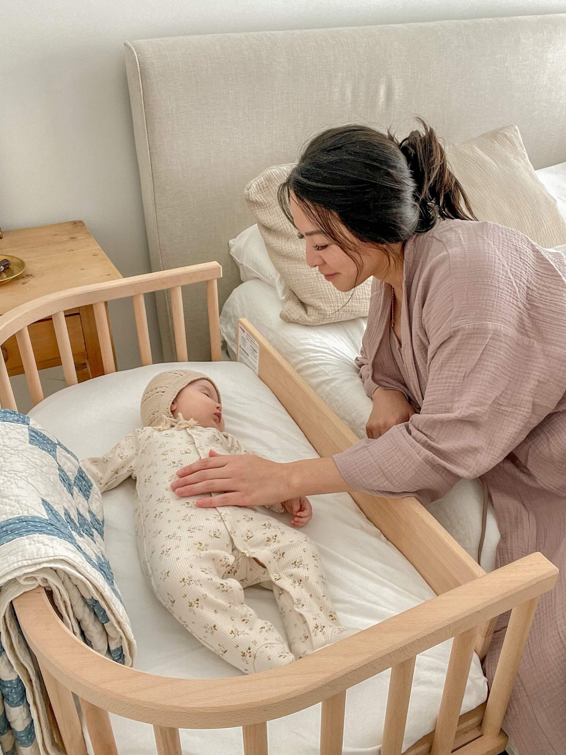 A woman in a robe gently places her hand on a baby lying in a wooden crib next to a bed, with a folded quilt draped over the crib rail.