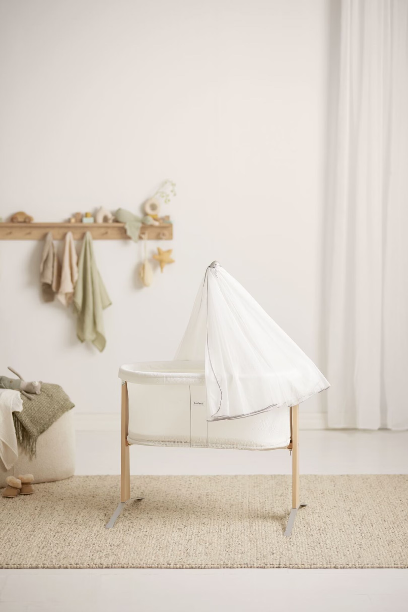 A white bassinet with a mesh canopy stands on a beige rug in a minimalist nursery with light wood accents and soft pastel decor.