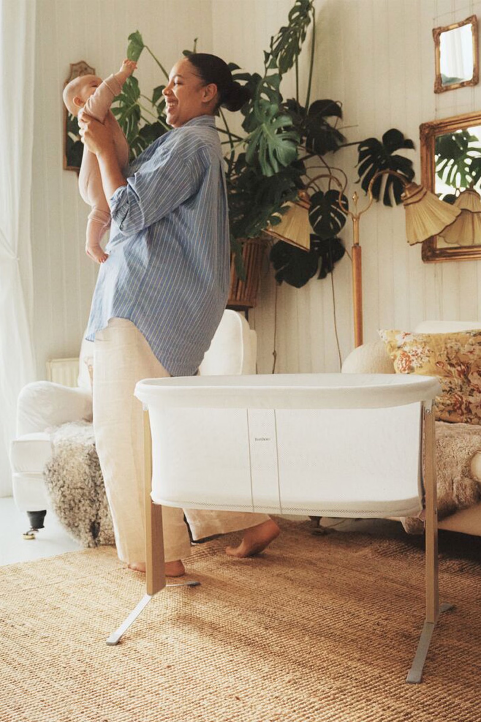A woman in a blue shirt and beige pants holds a baby in a cozy living room with a bassinet, plants, and cushioned furniture.