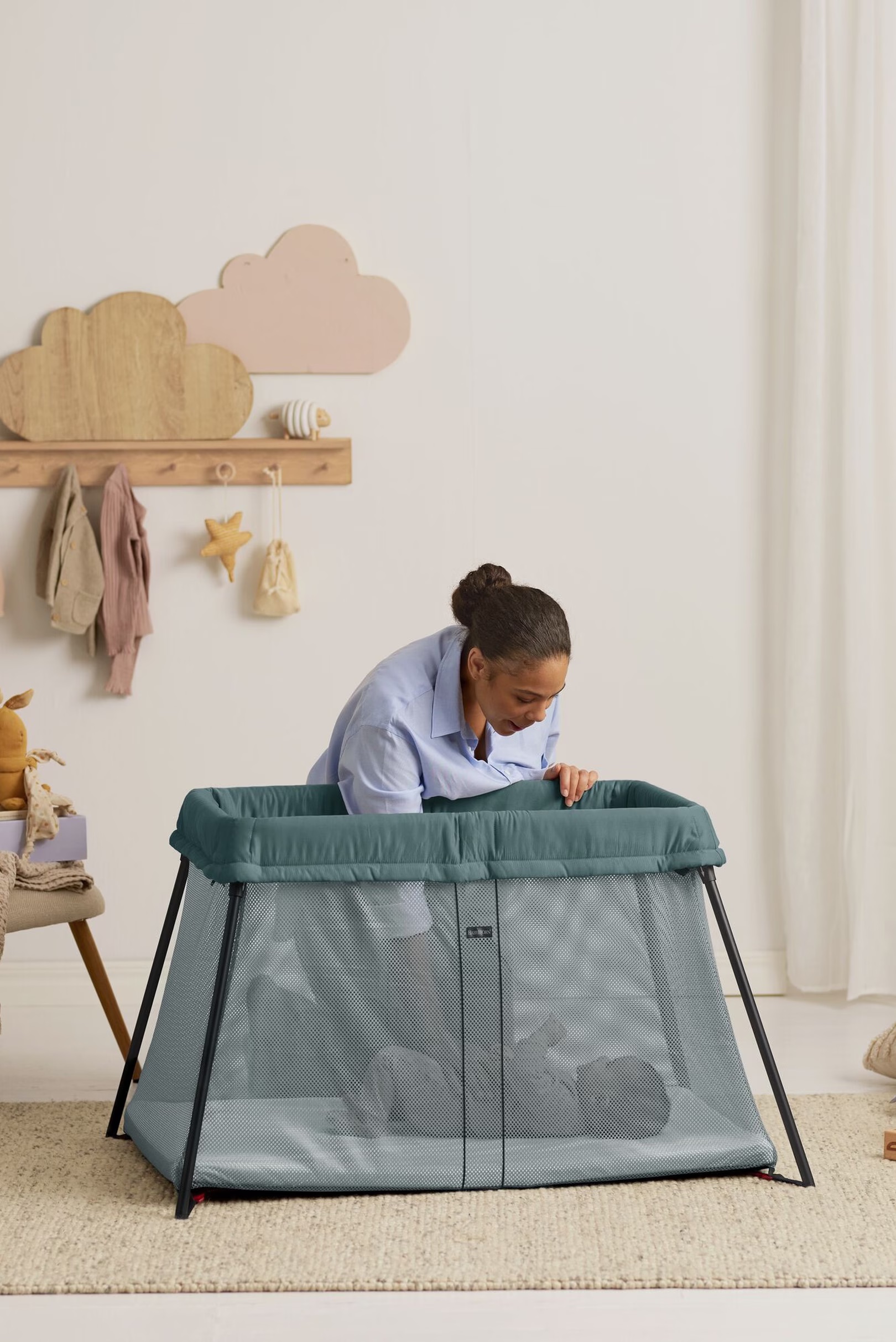 An adult leans over a teal playpen, looking at a baby lying inside. The room has a beige rug, toys, and wall decorations shaped like clouds.