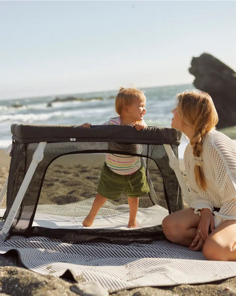 A woman sits on a blanket next to a baby standing inside a portable playpen on a sandy beach with the ocean in the background.