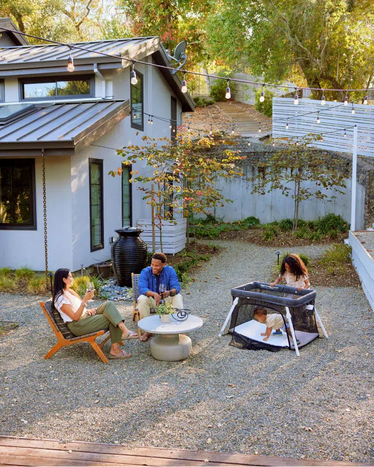 Three adults sit and talk in a modern backyard, while a child plays in a playpen. The scene is outdoors, next to a grey house with string lights overhead.