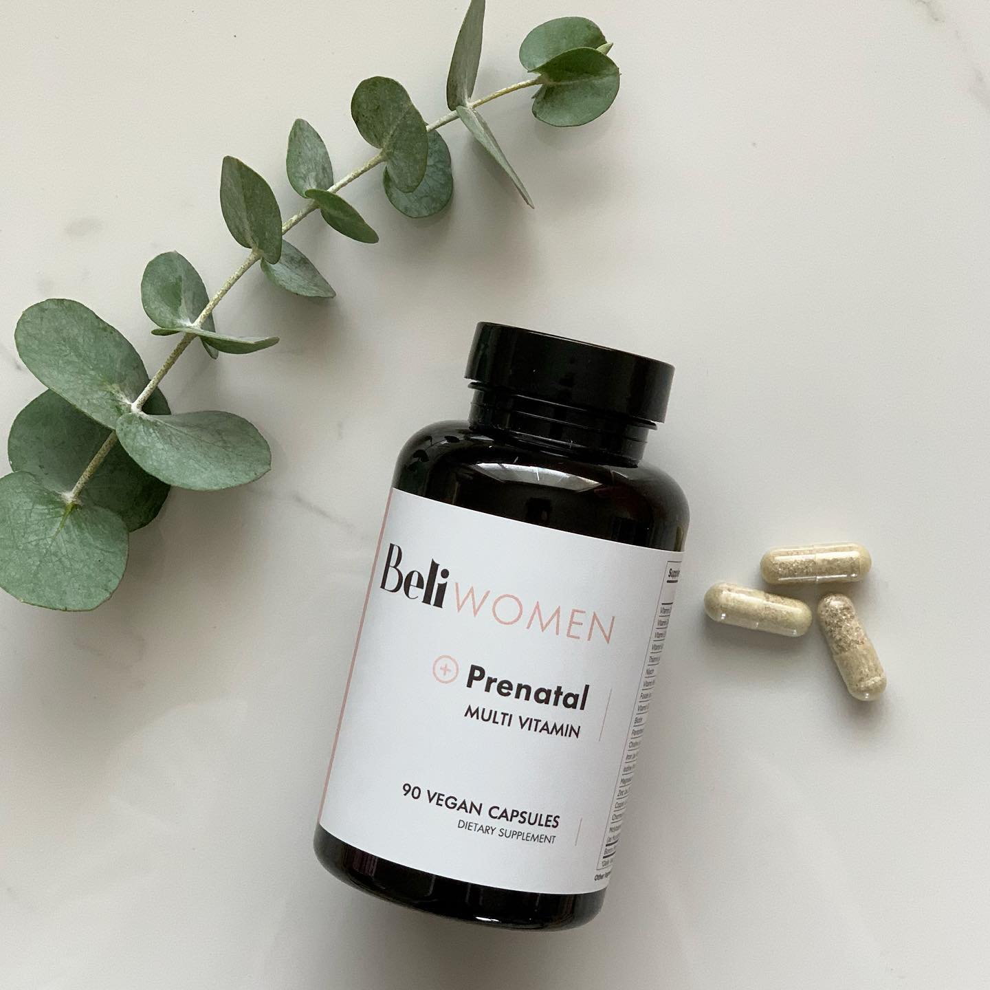 A bottle labeled "Beli WOMEN Prenatal Multivitamin" with three capsules beside it and a sprig of eucalyptus on a white surface.