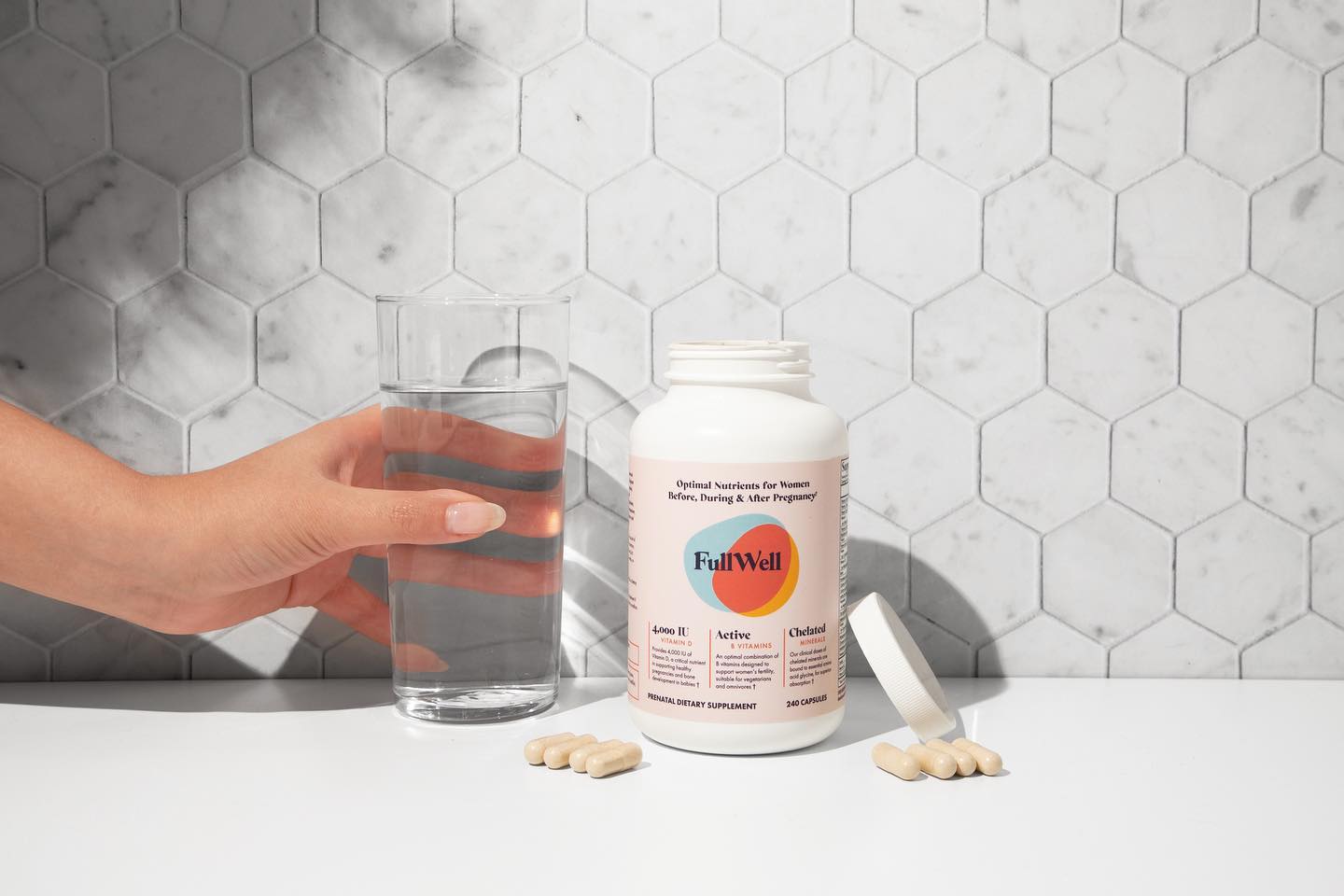 A hand holds a glass of water next to an open bottle of FullWell supplements with several capsules on a white surface against a hexagonal tile background.