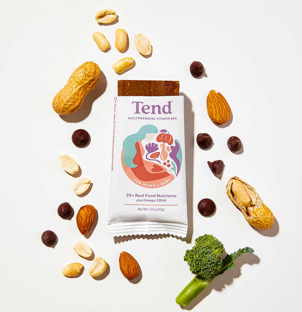 A Tend prenatal vitamin bar is surrounded by peanuts, almonds, chocolate chips, cashews, walnuts, and a small piece of broccoli on a white background.