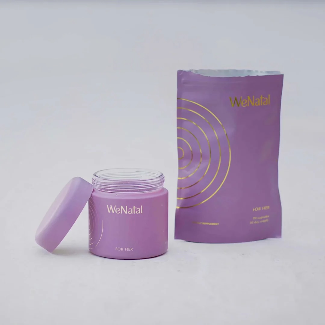 A purple WeNatal "For Her" supplement jar with its lid off sits next to a matching purple refill pouch with gold circular designs on a plain light background.