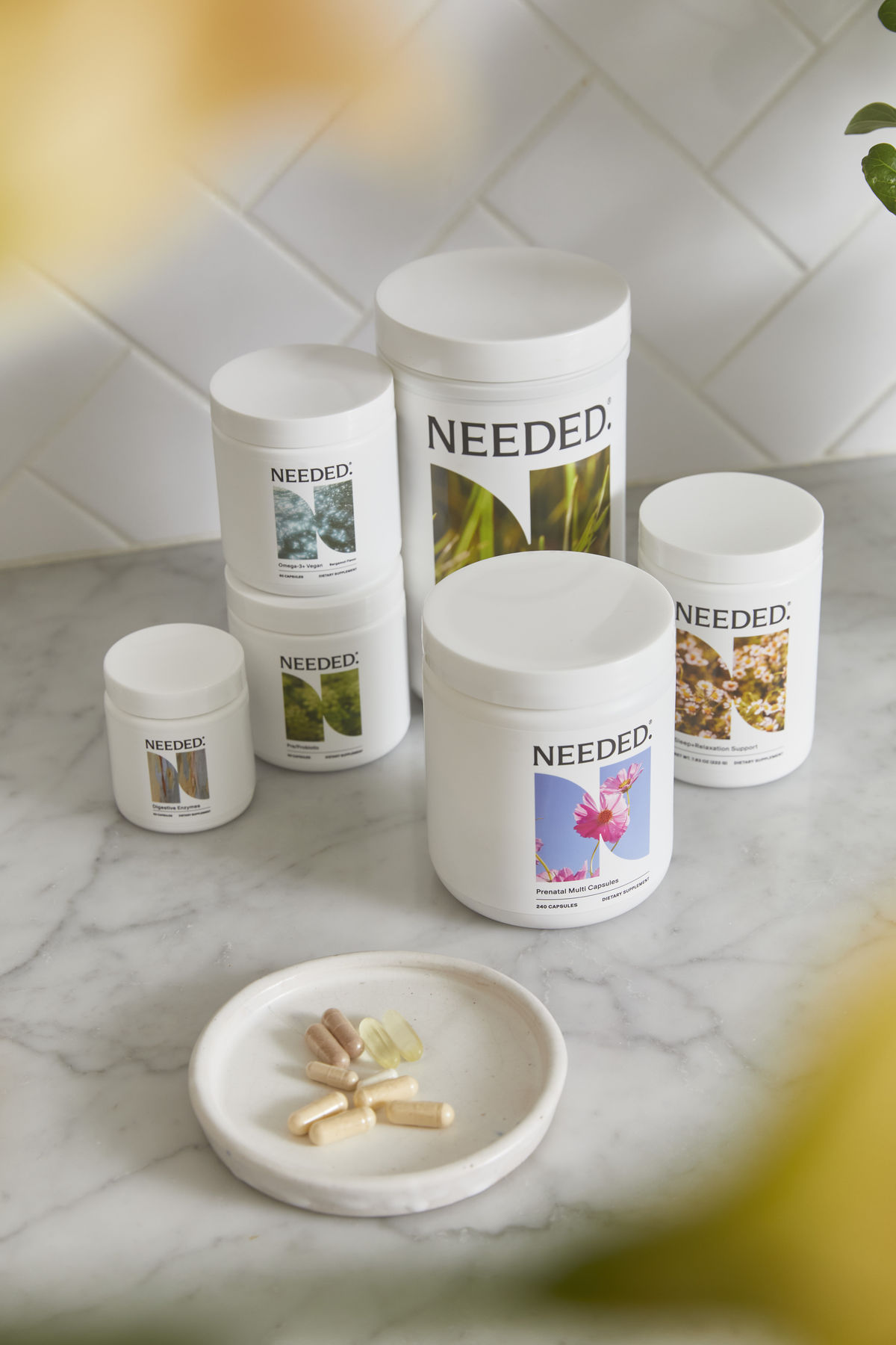 Various white supplement containers labeled "NEEDED." are arranged on a marble countertop, with a small dish holding different colored capsules and tablets in the foreground.