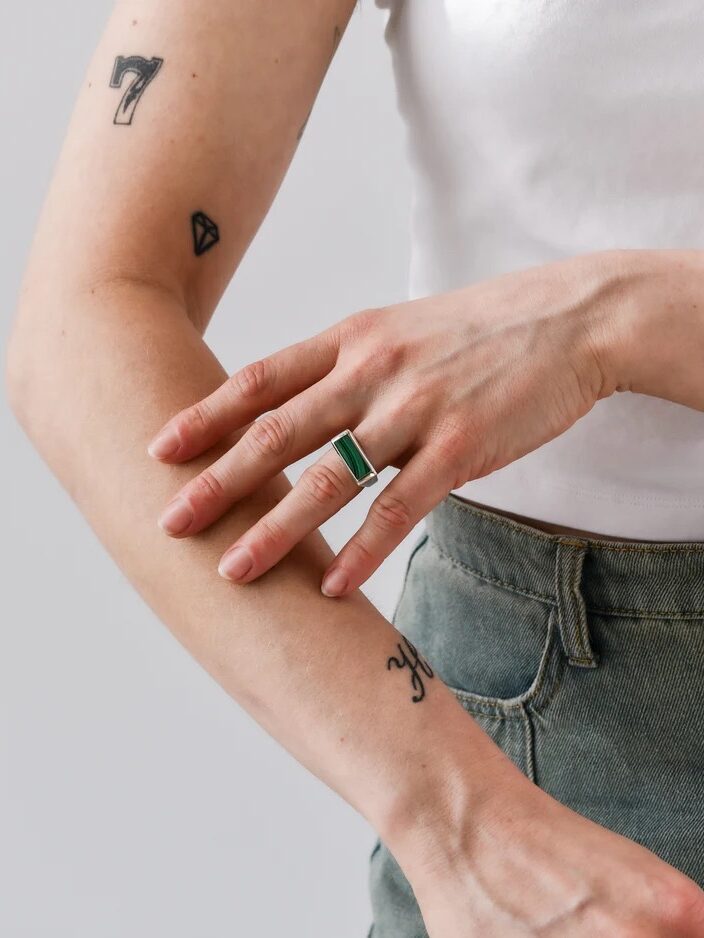 A person wearing a white shirt and green pants touches their forearm, showing tattoos and a rectangular green stone ring on their finger.