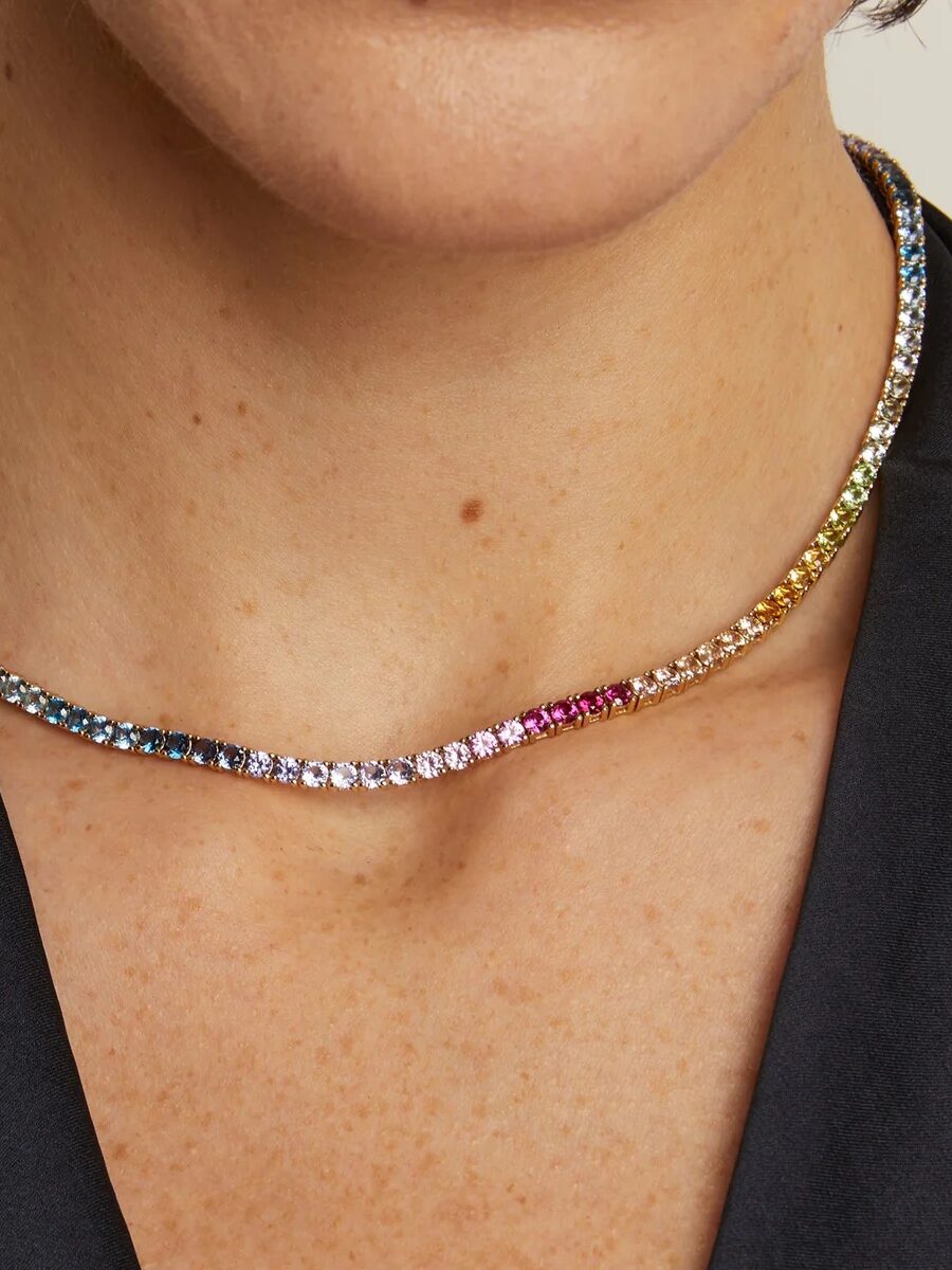 A close-up of a person wearing a black top and a multicolored gemstone tennis necklace around their neck.