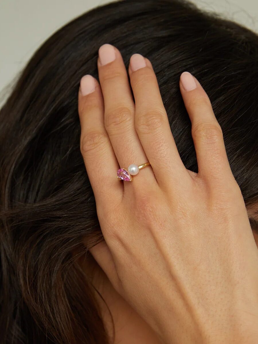A hand with light pink nail polish rests on dark hair, wearing a gold ring featuring a pink gemstone and a white pearl.