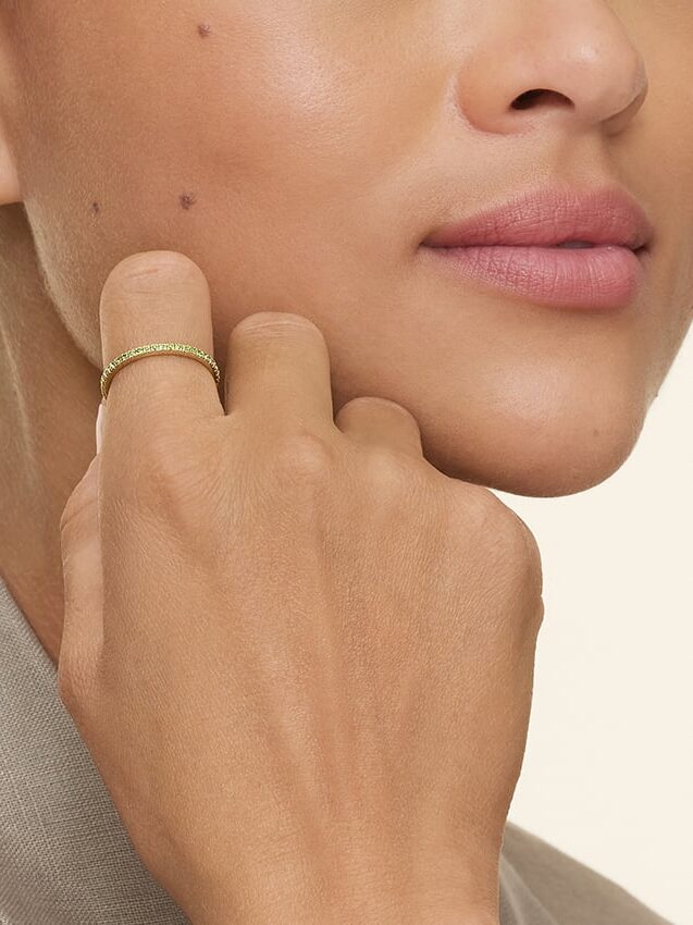 Close-up of a woman’s face and hand, showing a gold ring with small green stones on her finger and a stud earring in her ear. She is wearing a taupe-colored top.