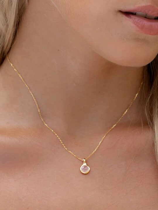 A woman wears a delicate gold chain necklace with a small, round pendant featuring a light-colored stone.