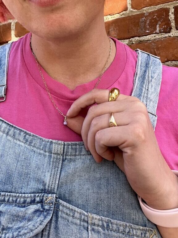 A person wearing a pink shirt and denim overalls touches a necklace, showing gold rings on their fingers, with a brick wall in the background.