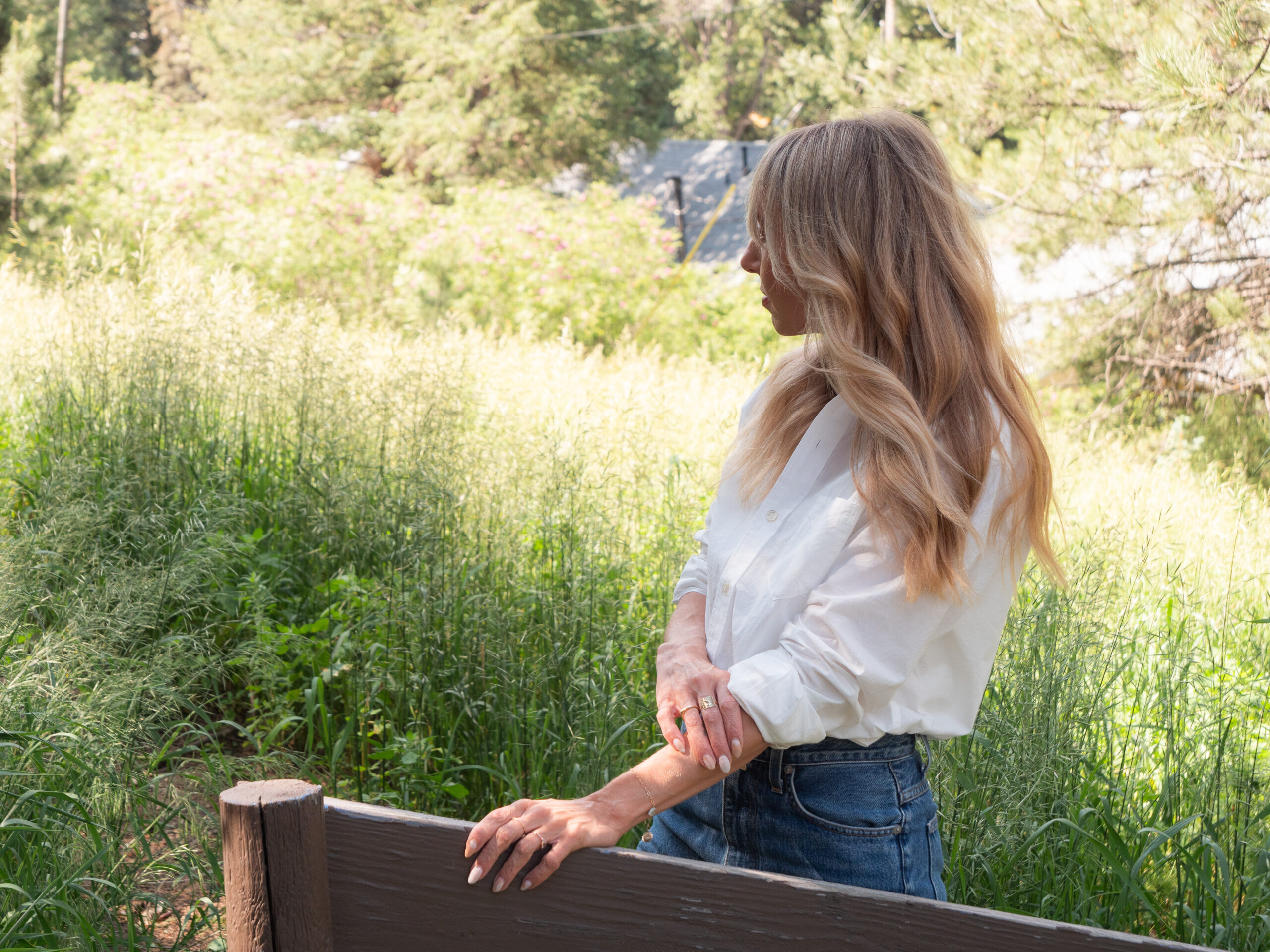 A woman with long blonde hair, wearing a white shirt and blue jeans, stands outdoors by a wooden fence, looking to her right with greenery and trees in the background.