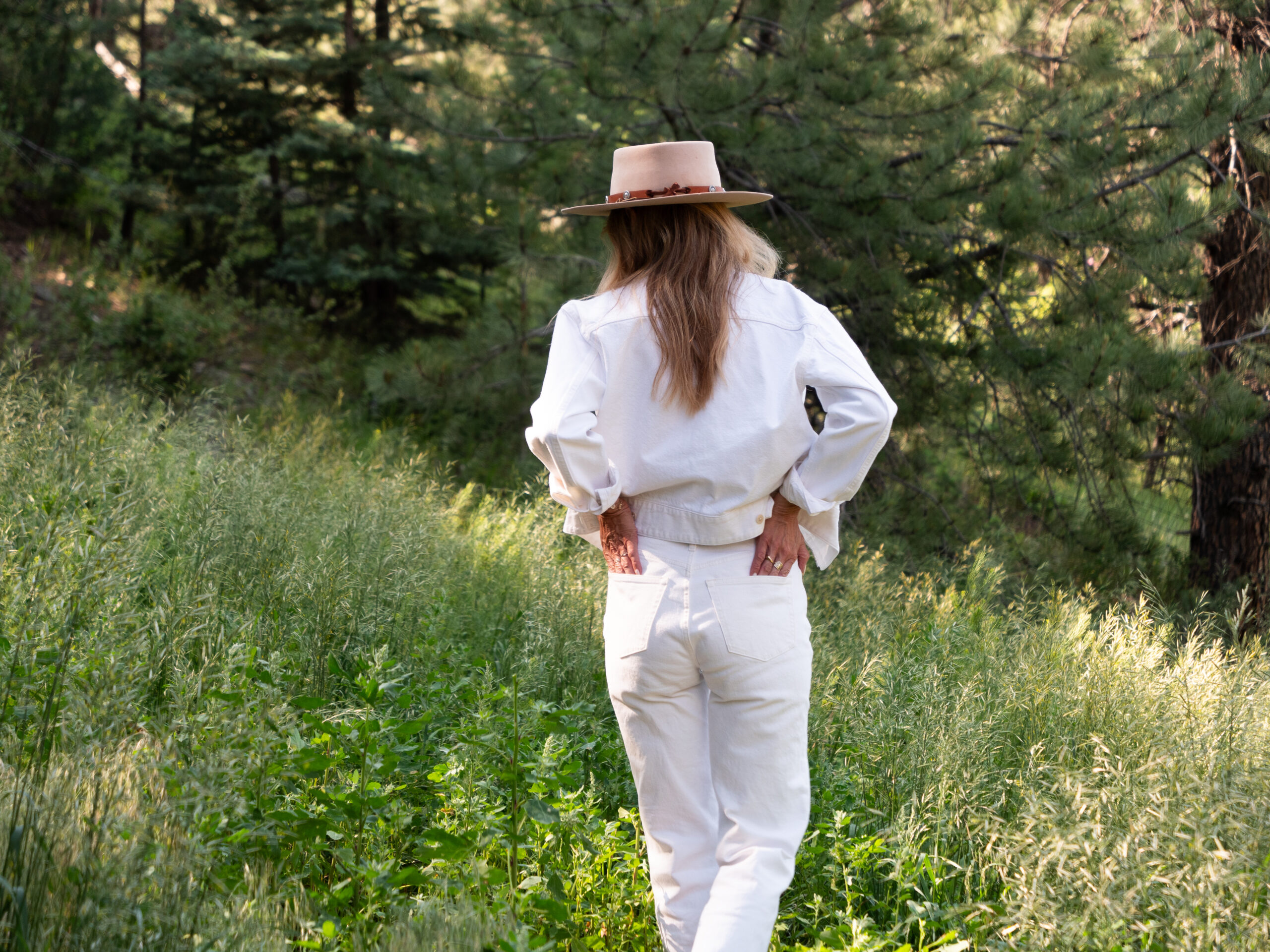 A person wearing a wide-brimmed hat and white outfit stands with hands on hips, facing away, in a grassy area surrounded by trees.
