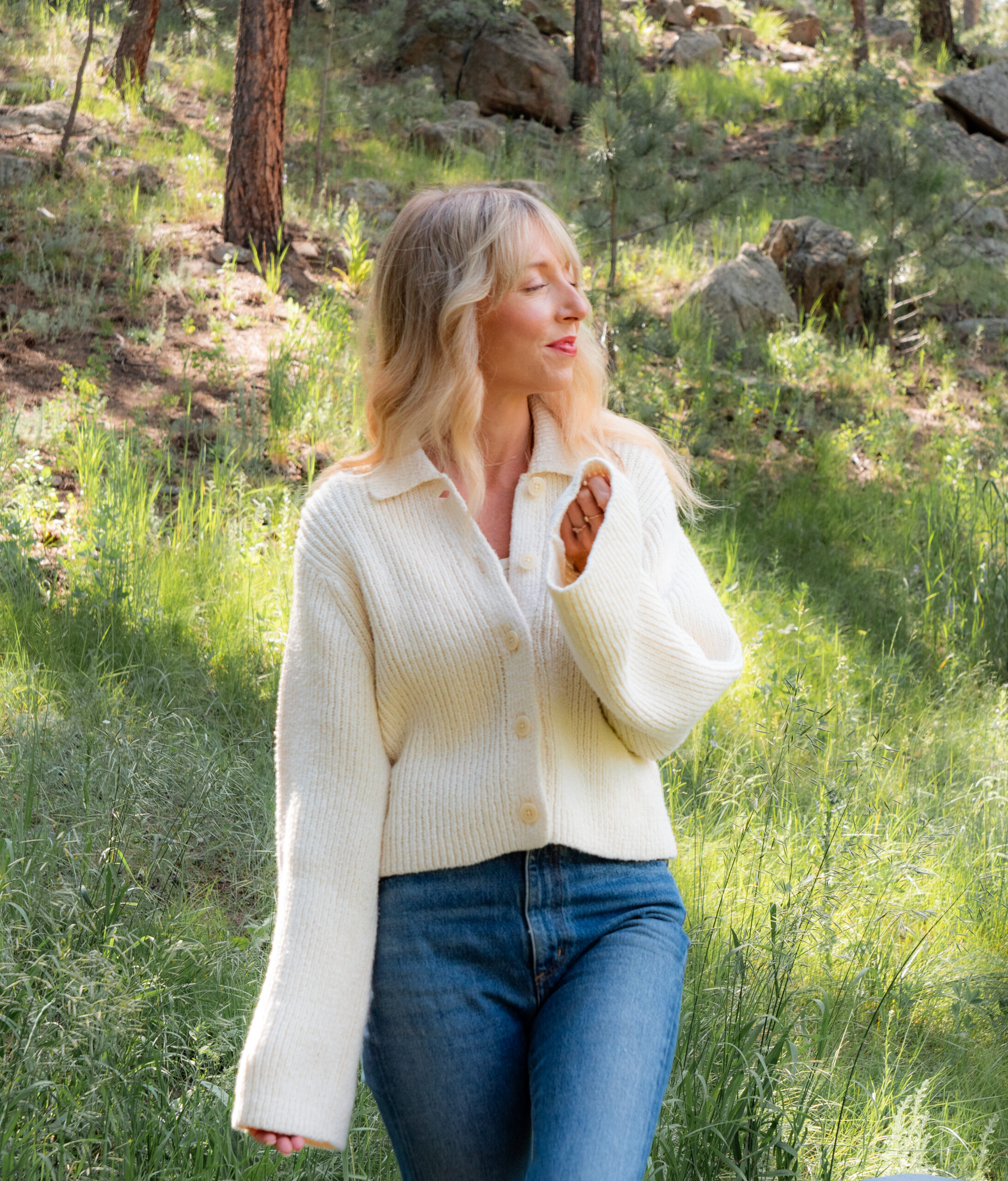 A person with blonde hair wearing a white sweater and blue jeans stands outdoors in a sunlit wooded area with green grass and trees.