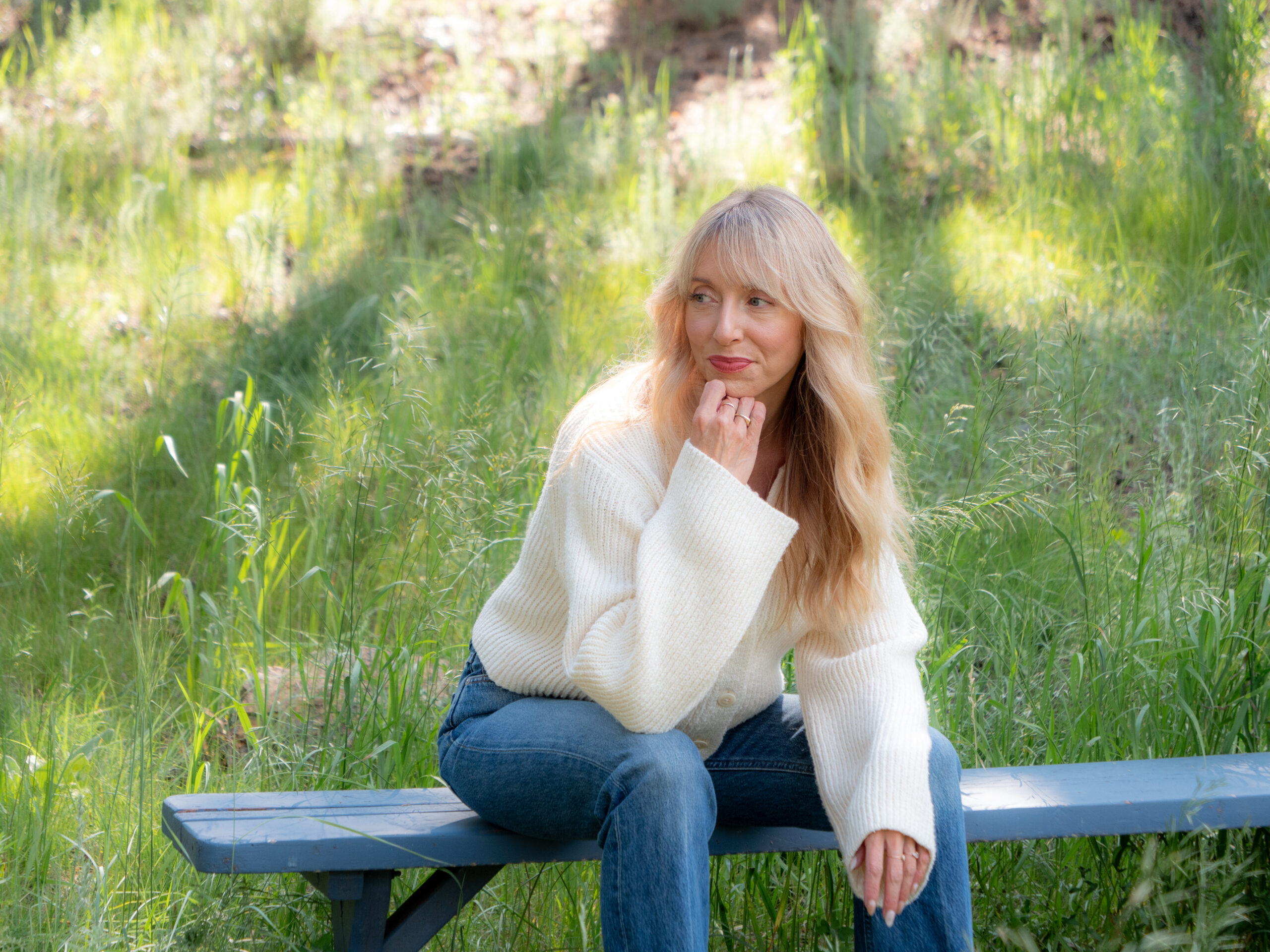 A woman with long blonde hair wearing a white sweater and jeans sits on a bench outdoors, surrounded by green grass and sunlight.