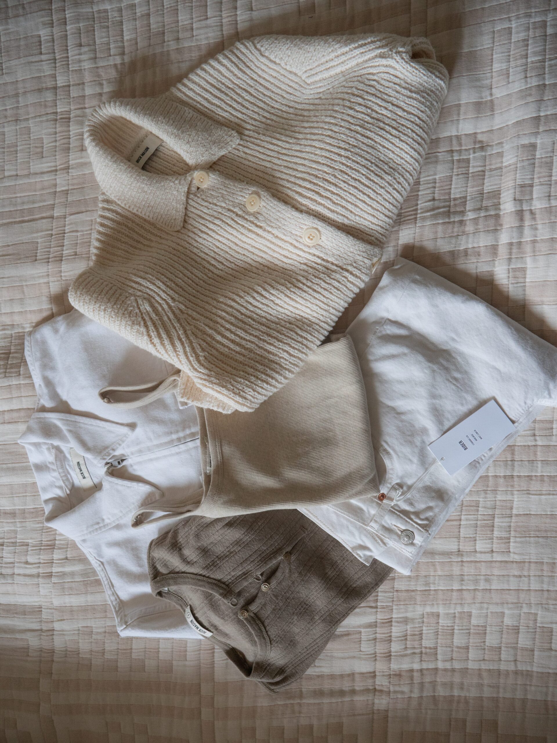 A neatly arranged set of beige and white clothing items, including shirts, pants, and a knit sweater, displayed on a textured quilted surface.